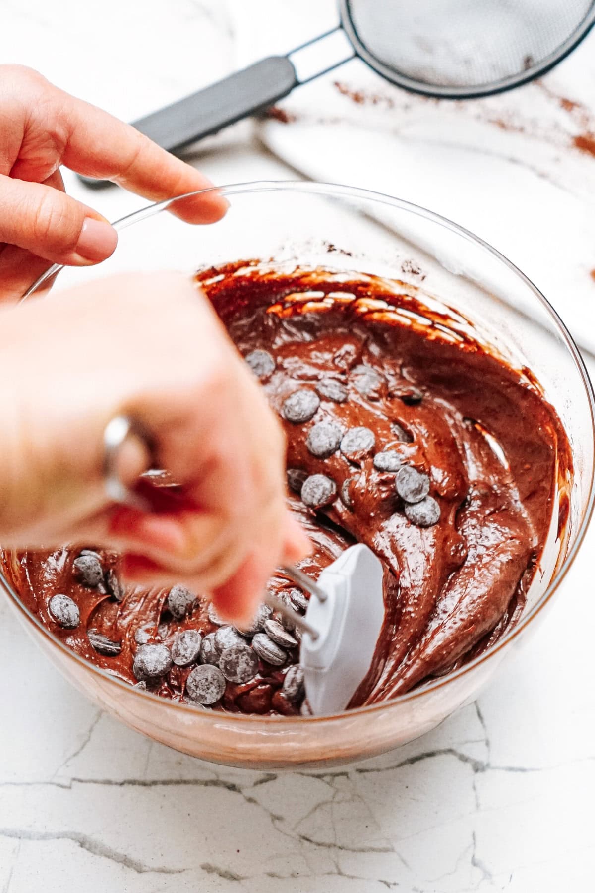 Hand stirring chocolate chips into brownie batter in a glass bowl with a spatula. A sifter is visible in the background on a marble countertop.