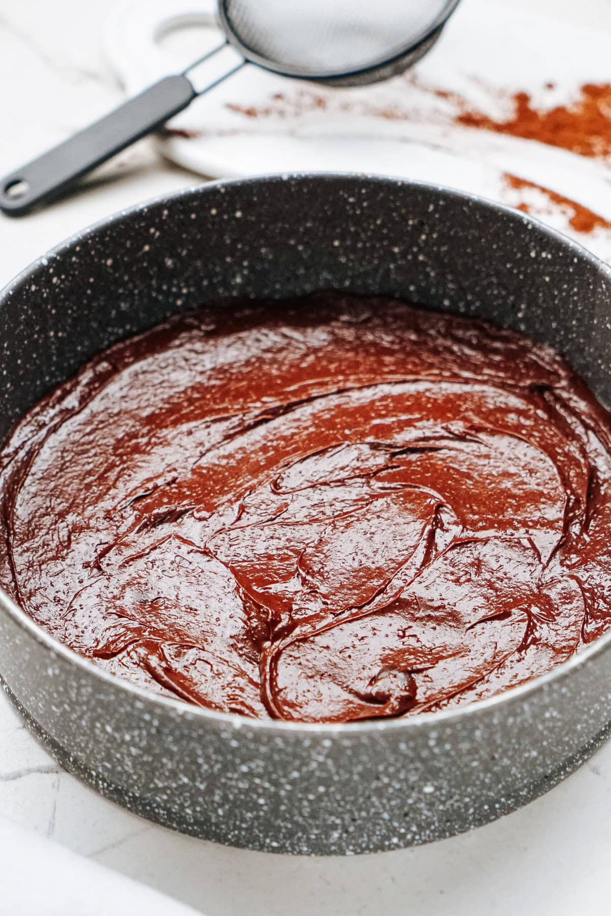 A round baking pan filled with smooth chocolate cake batter. A sifter and cocoa powder are visible in the background.