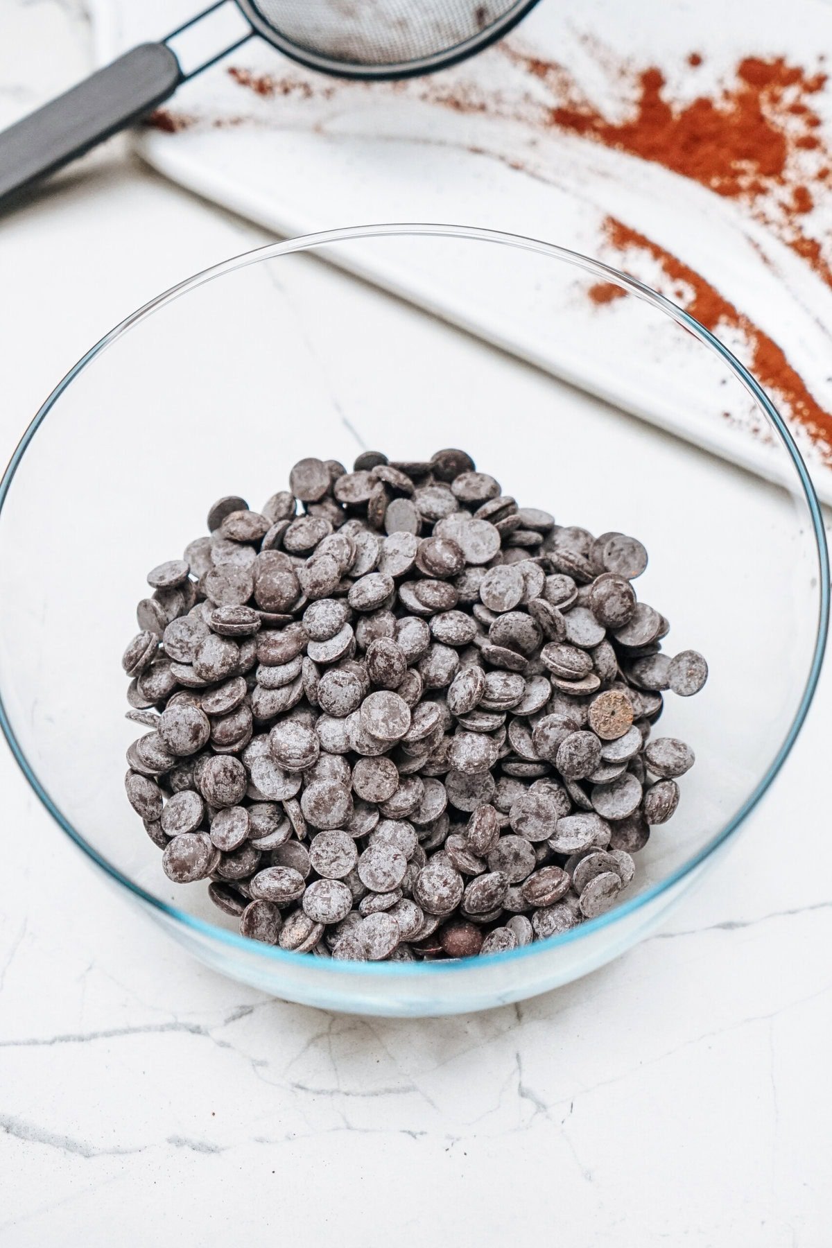 A glass bowl filled with chocolate chips on a marble countertop. A sieve and cocoa powder are partially visible in the background.