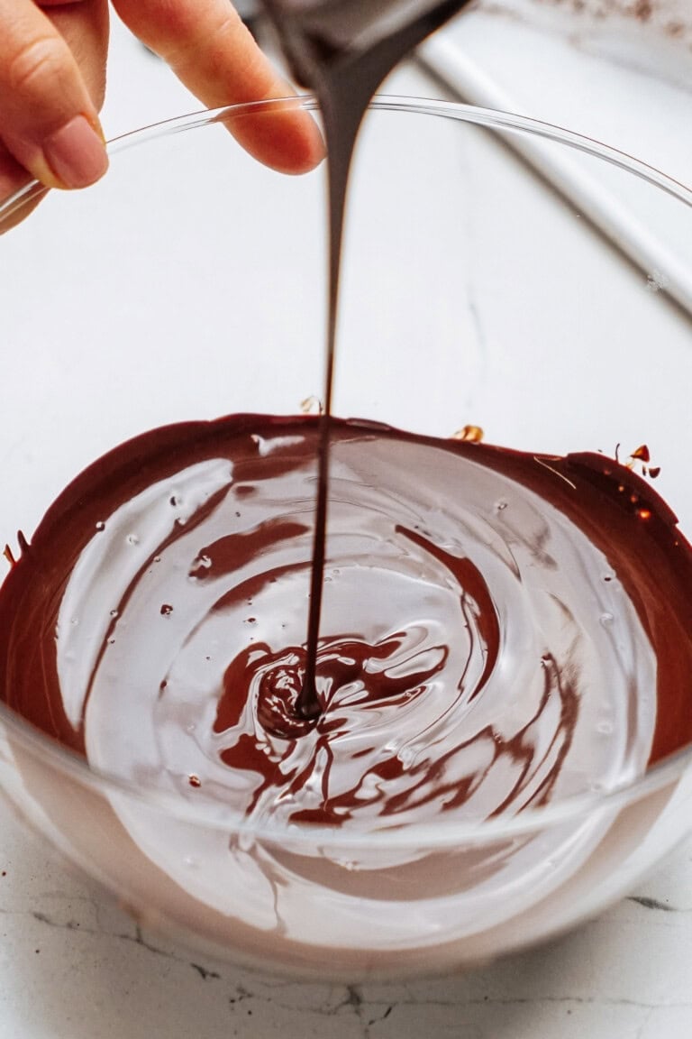 Melted chocolate is being poured into a glass bowl, creating swirls on the surface.