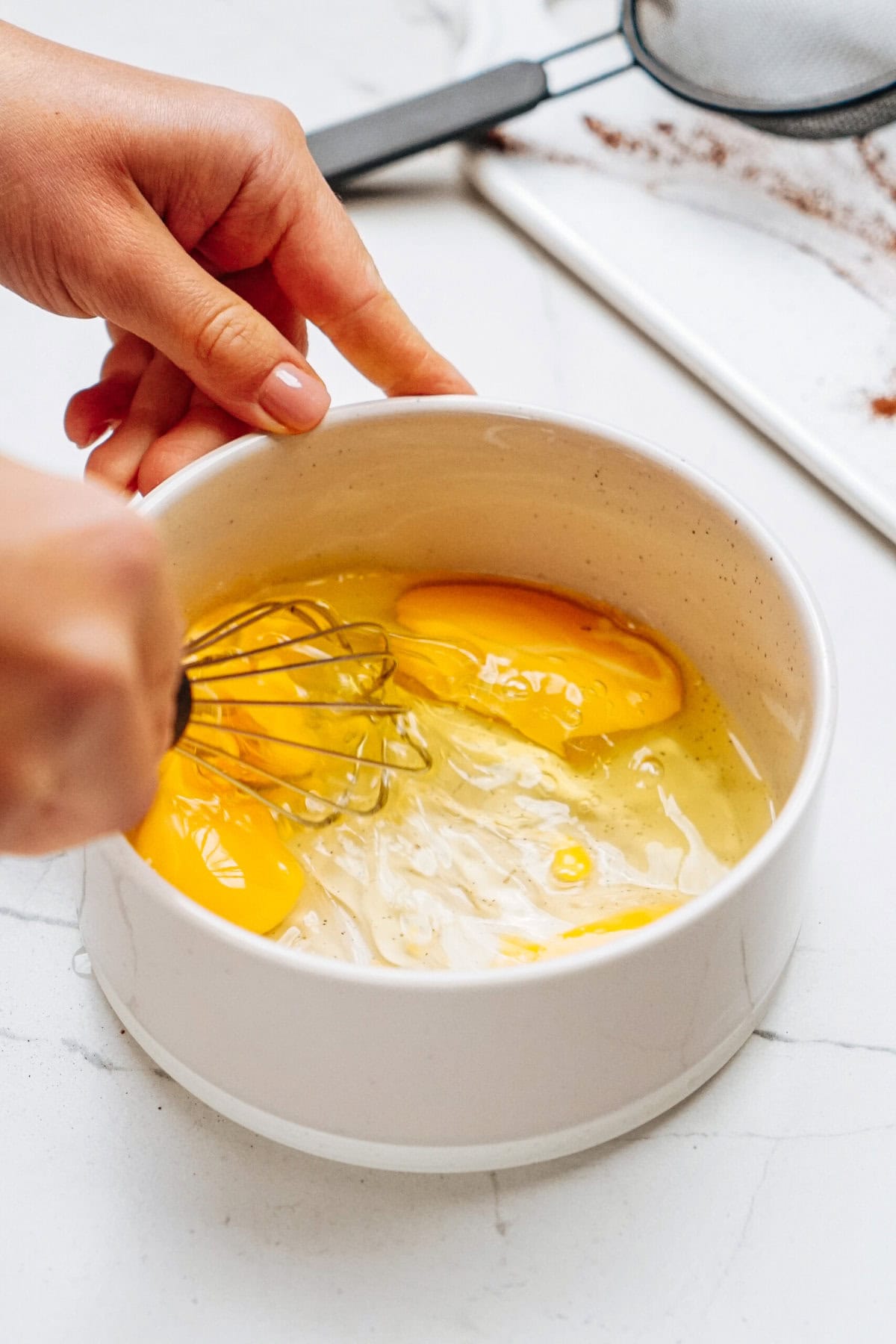 Hands whisking eggs in a white bowl on a kitchen counter.