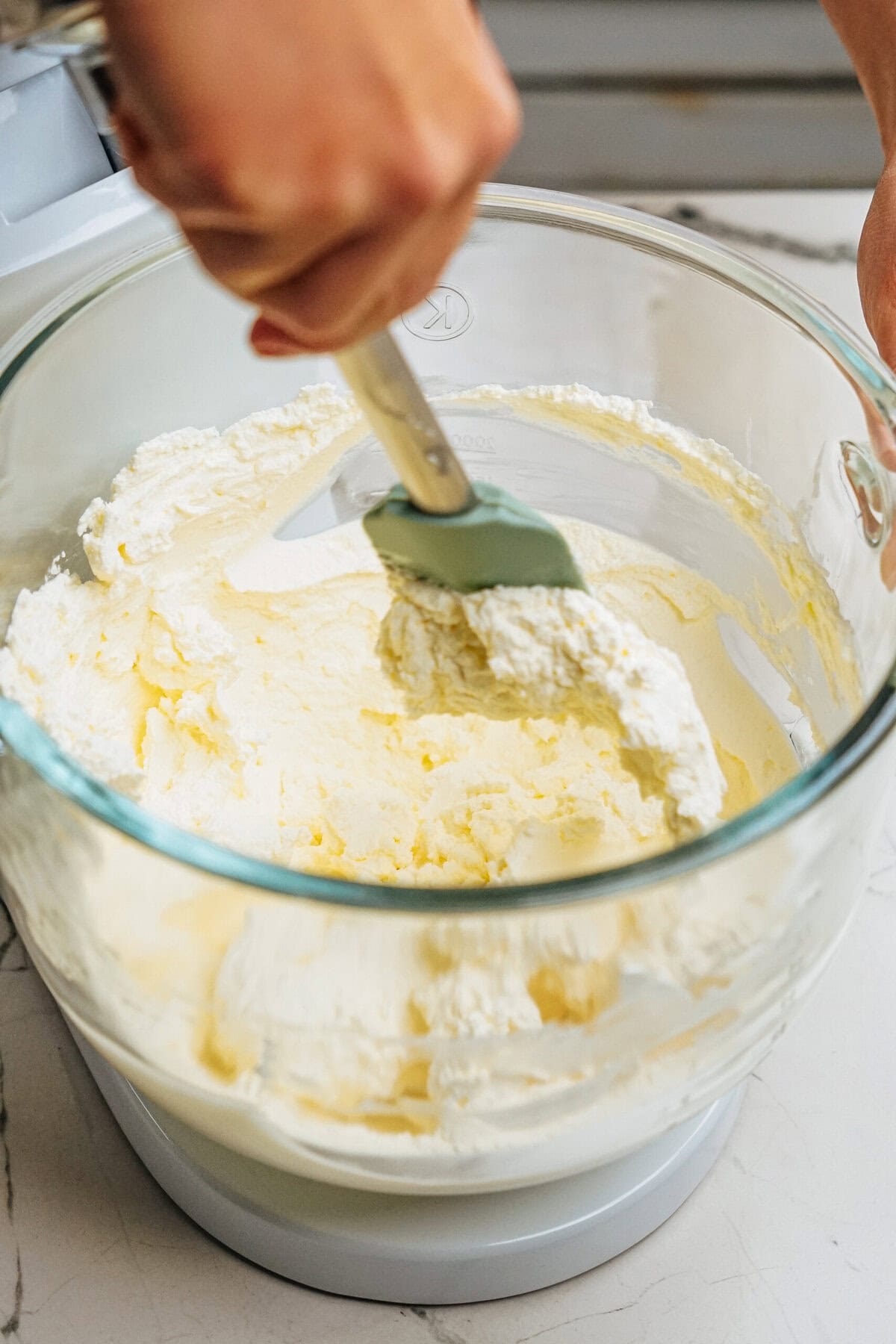 A person uses a spatula to mix a creamy batter in a clear glass mixing bowl.