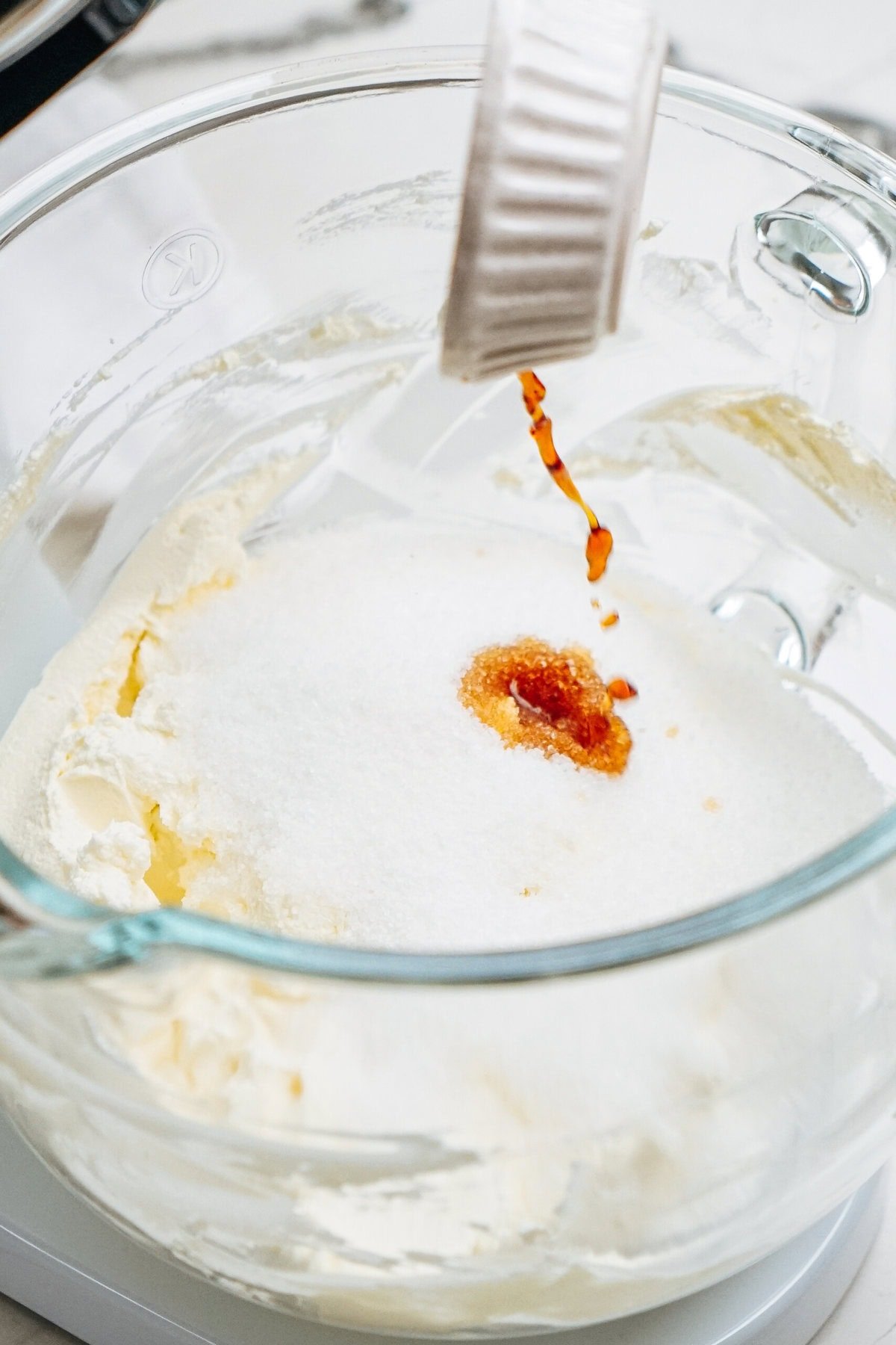 A clear bowl with cream and sugar is being mixed. Brown liquid, possibly vanilla extract, is being poured in from a white container.