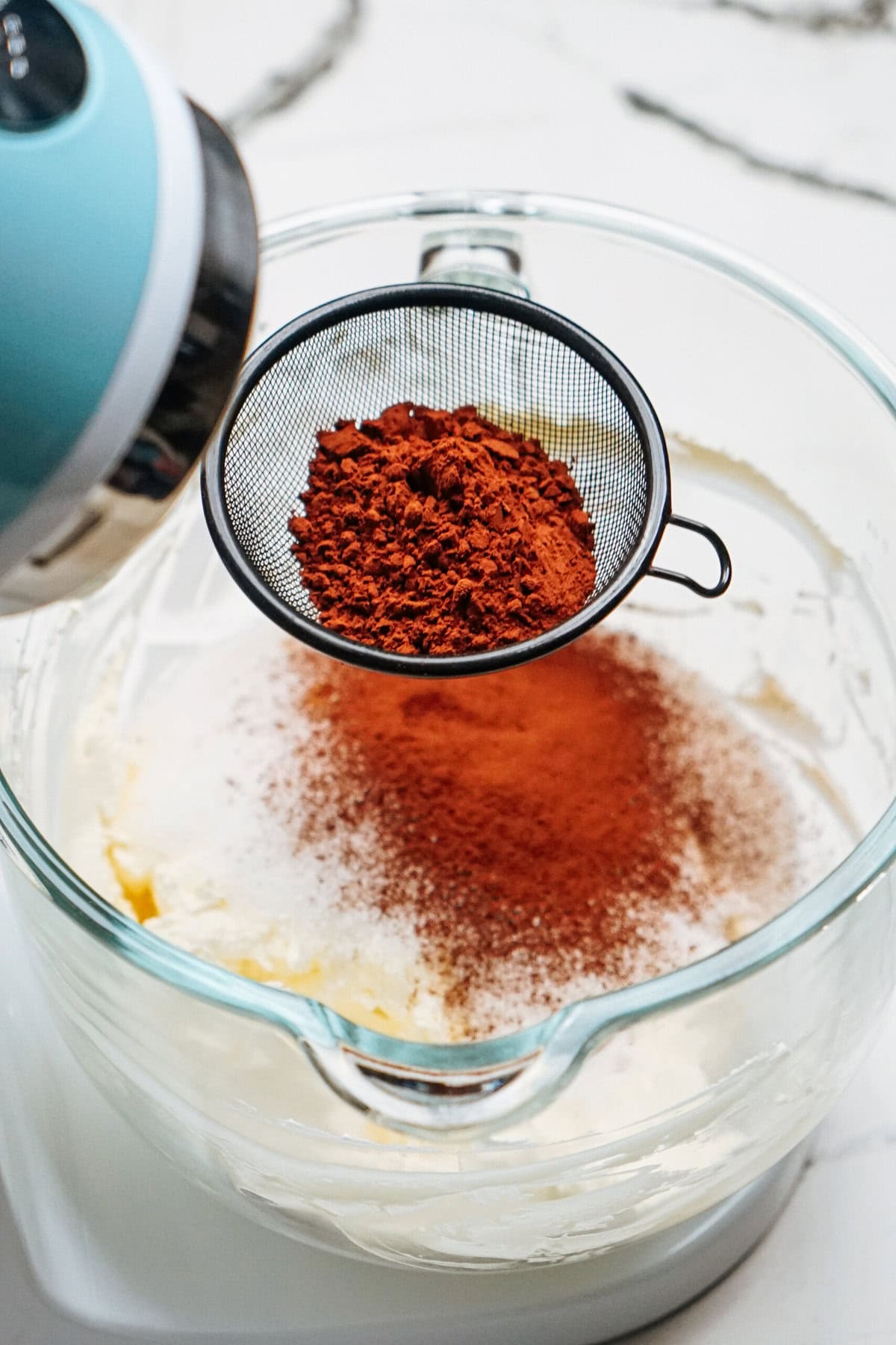 A sifter with cocoa powder is held above a glass mixing bowl containing flour, butter, and sugar.