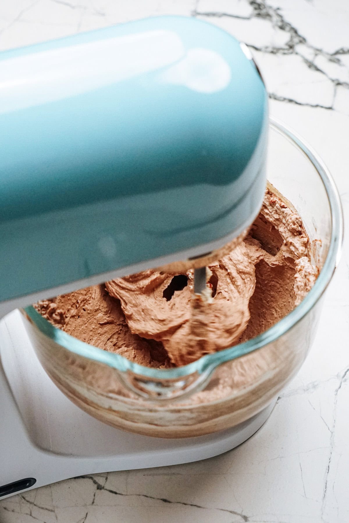 A blue stand mixer with a glass bowl containing chocolate batter on a marble countertop.