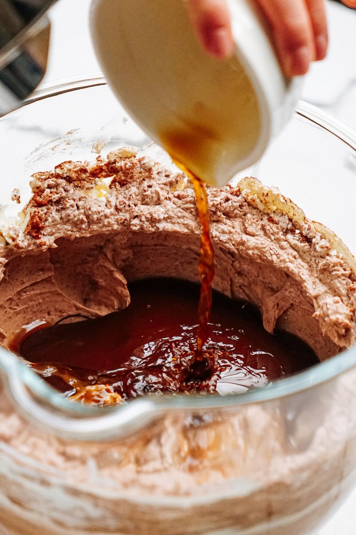 A hand pours liquid into a mixing bowl filled with chocolate batter.