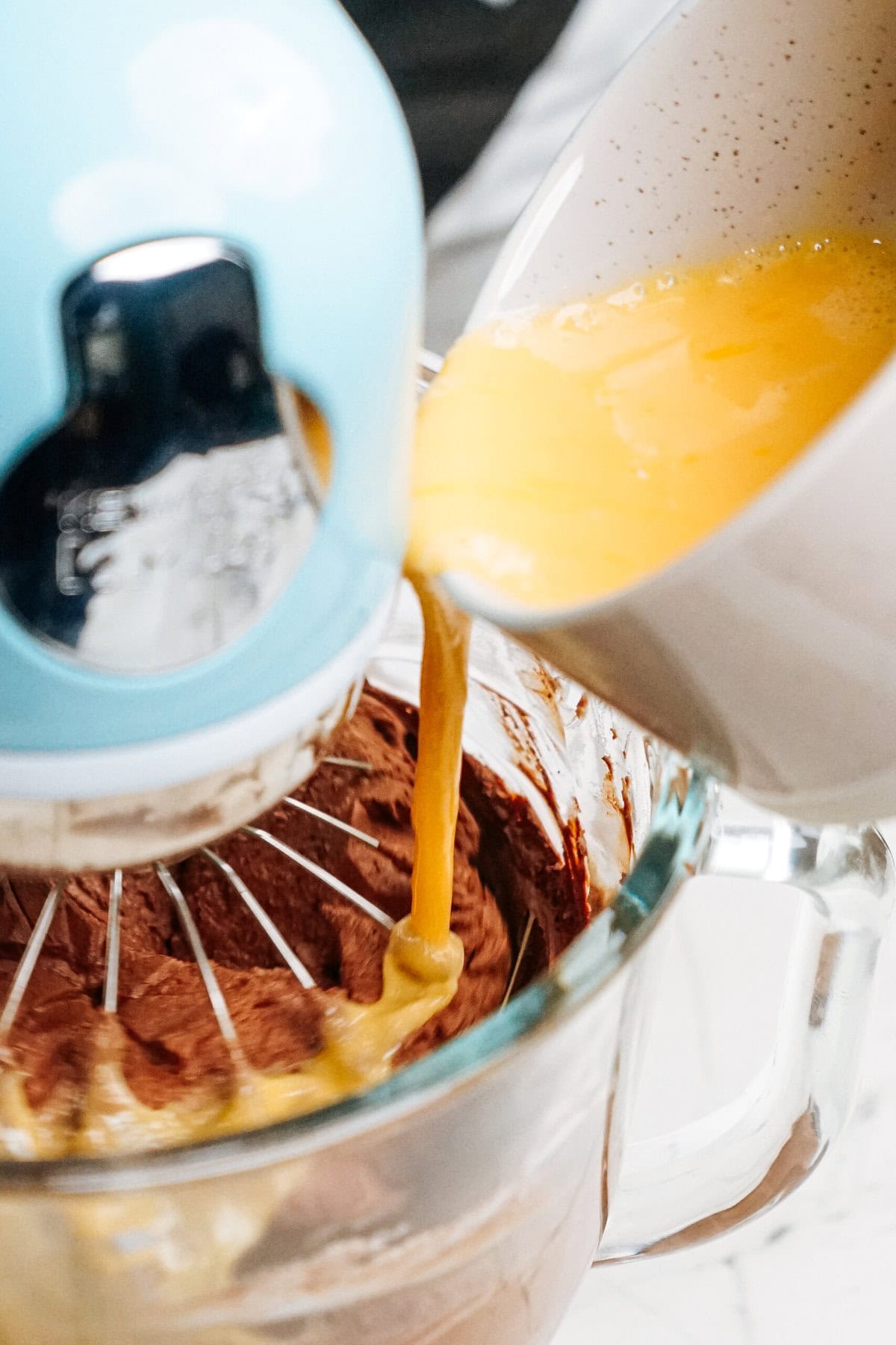 A person pours a creamy yellow liquid into a mixing bowl with chocolate batter while a blue stand mixer stirs.