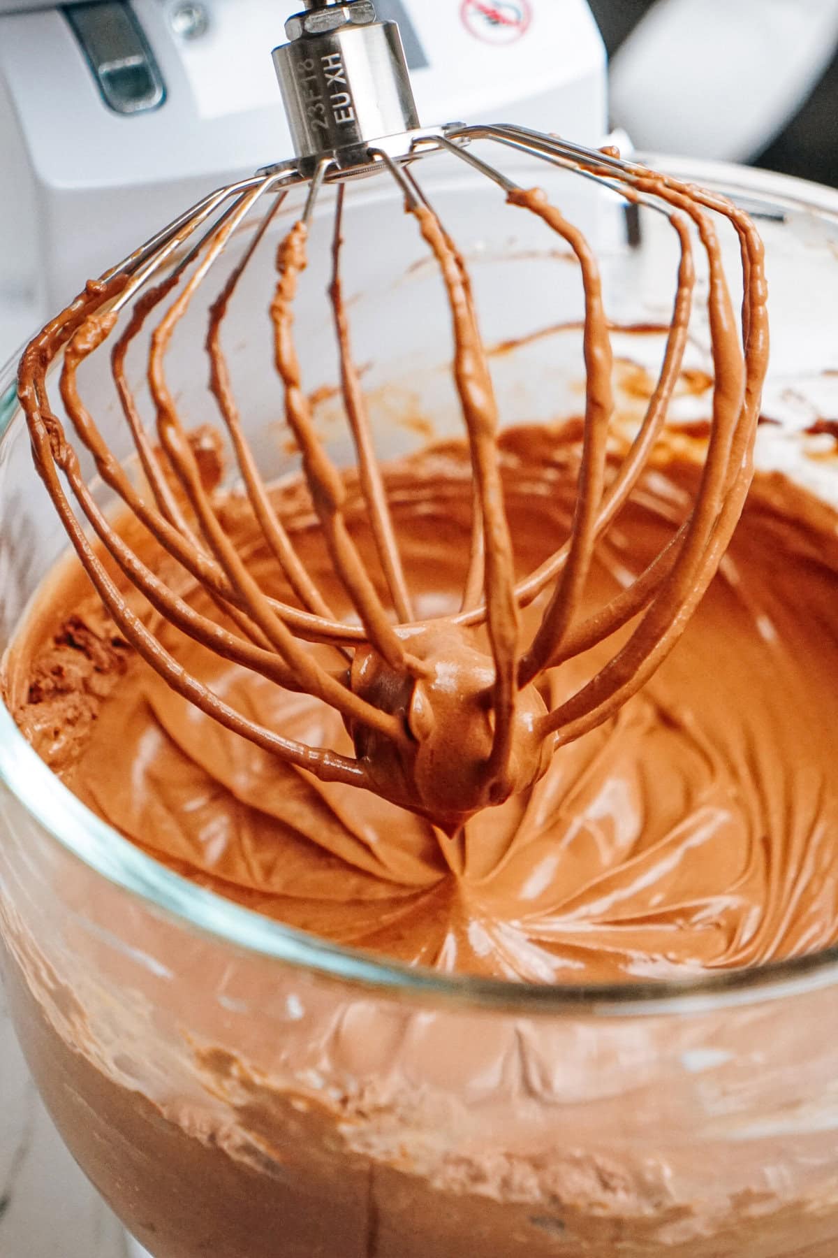 Close-up of a mixer whisk blending smooth chocolate batter in a glass bowl.