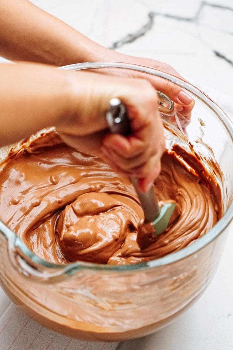 A person stirs chocolate cake batter with a spatula in a glass mixing bowl.