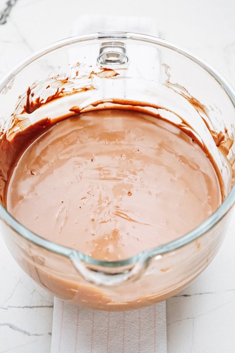 A glass bowl containing smooth, light brown chocolate batter on a white marble surface.