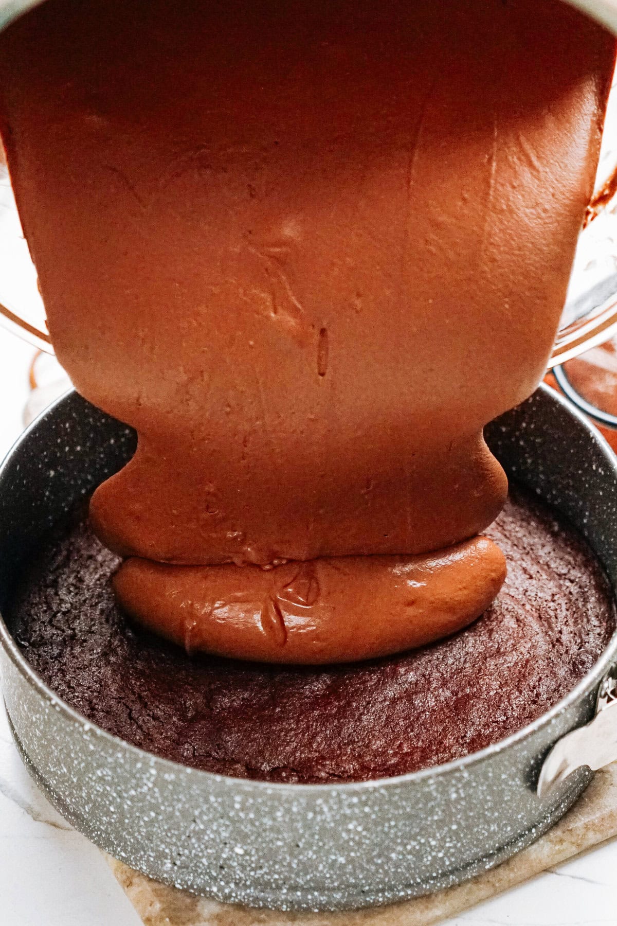 Chocolate batter being poured into a gray round cake pan on top of already baked cake layer.