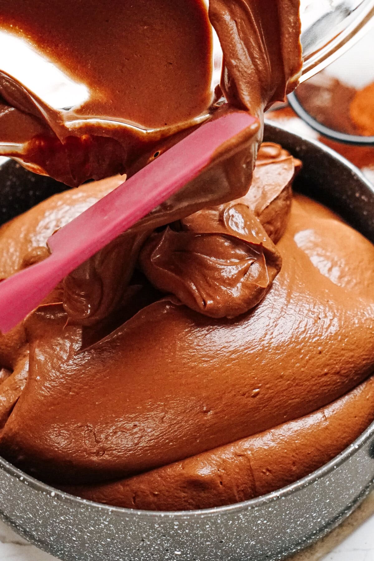 Chocolate cake batter being poured from a bowl into a baking pan, with a pink spatula assisting in spreading the mixture evenly.