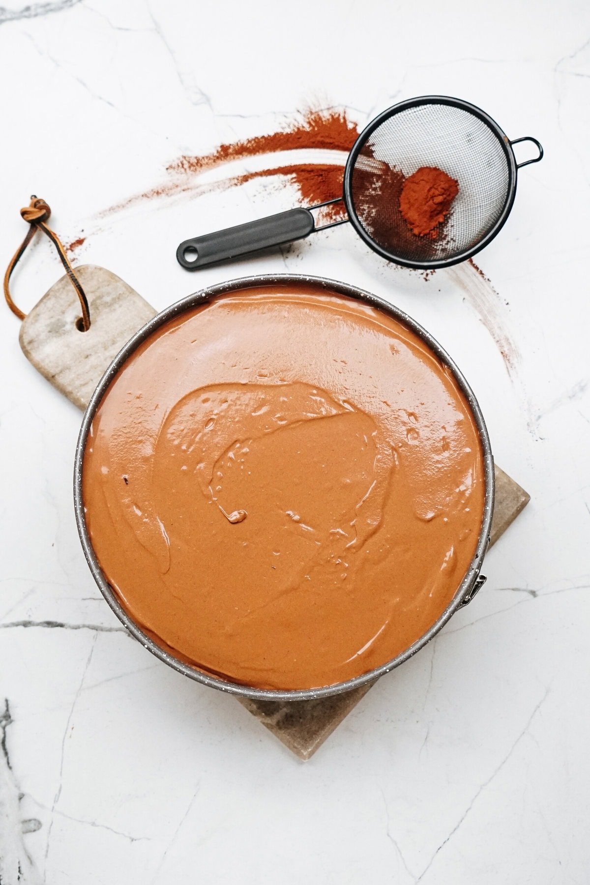 A round pan of smooth, creamy chocolate batter on a marble surface next to a sieve with cocoa powder.