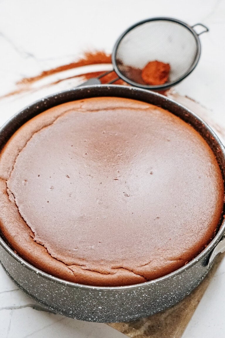 A round chocolate cake in a baking pan on a marble surface, with a mesh strainer holding cocoa powder nearby.