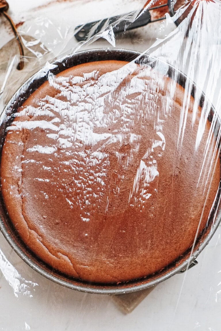 A freshly baked cake covered with plastic wrap in a round baking pan, resting on a wooden surface.