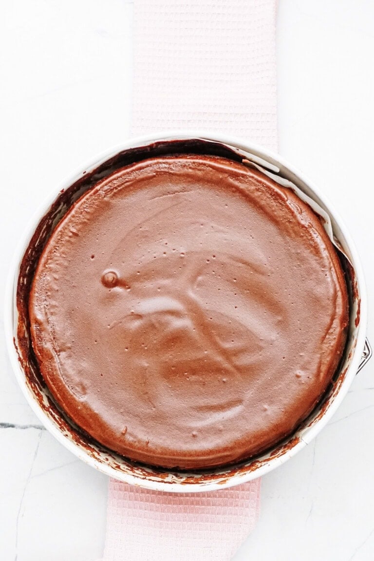 A chocolate cake in a round pan with a smooth, glossy surface, placed on a light pink textured cloth, on a white marble background.