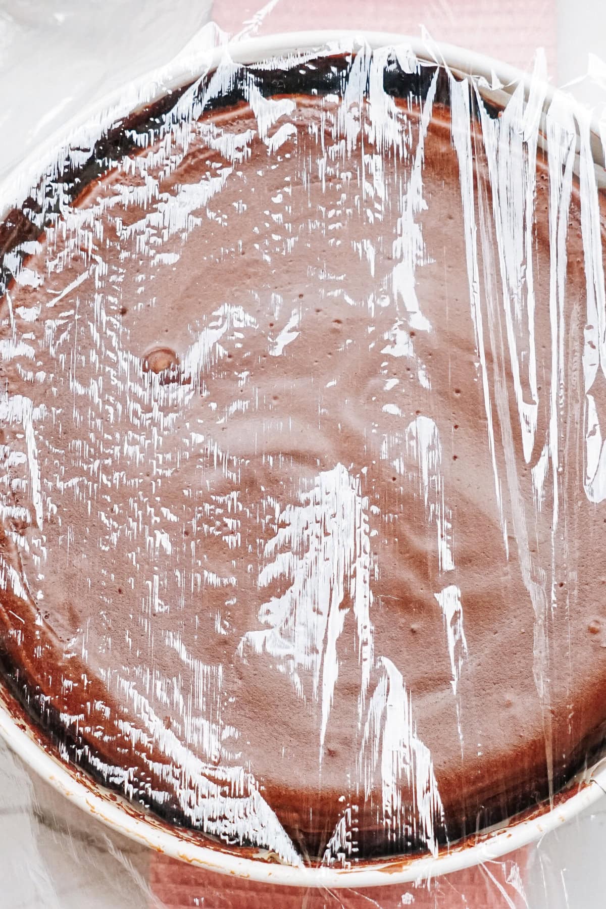 Chocolate cake covered with plastic wrap in a round dish.