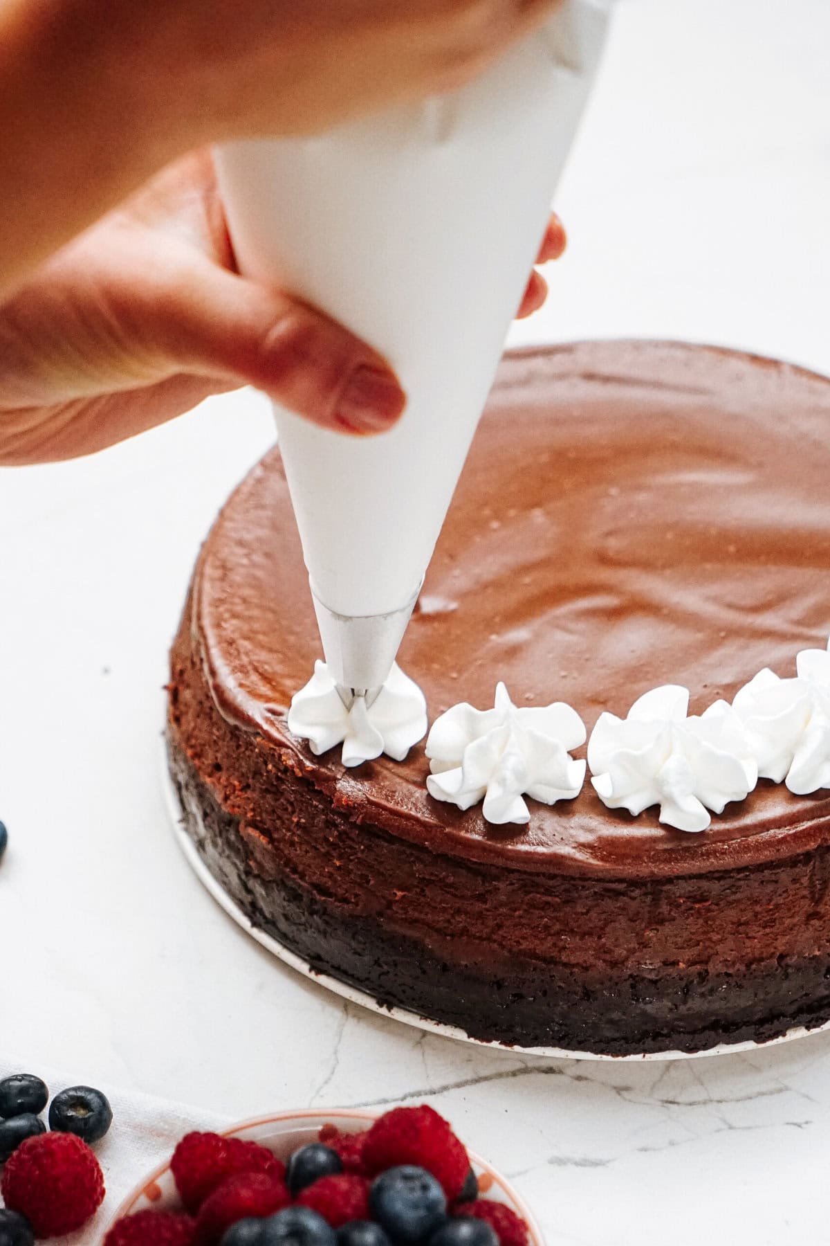 Person decorating a chocolate cake with white frosting using a piping bag. Bowl of mixed berries in the foreground.