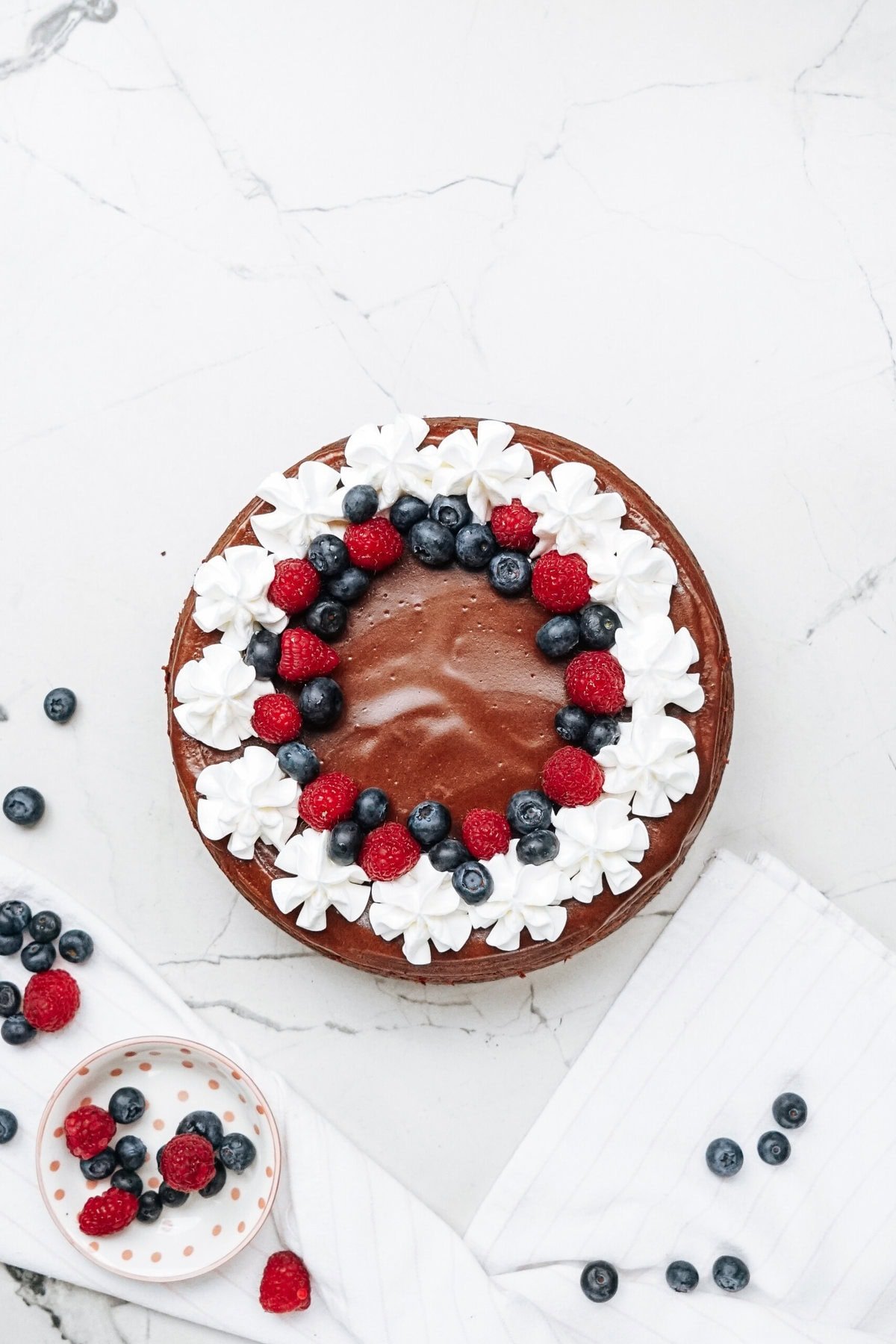 Chocolate cake with whipped cream, blueberries, and raspberries on top, set on a marble surface with scattered berries and a striped napkin nearby.