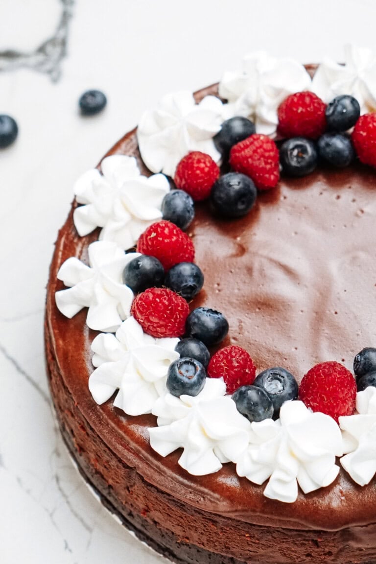 Chocolate cheesecake topped with whipped cream, raspberries, and blueberries on a marble surface.