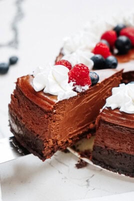 A sliced chocolate cheesecake topped with whipped cream, raspberries, and blueberries on a marble surface.