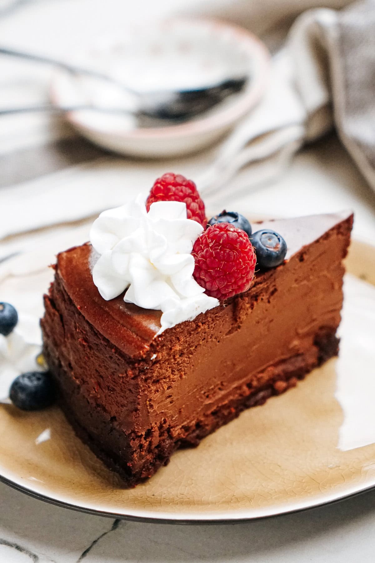 A slice of chocolate cheesecake topped with whipped cream, raspberries, and blueberries on a beige plate.