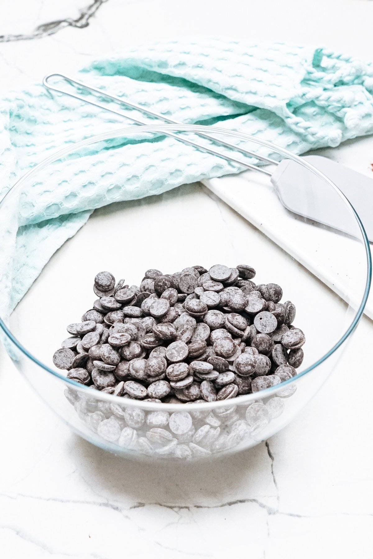 A glass bowl filled with dark chocolate chips sits on a white marble surface, near a light blue cloth and a spatula.