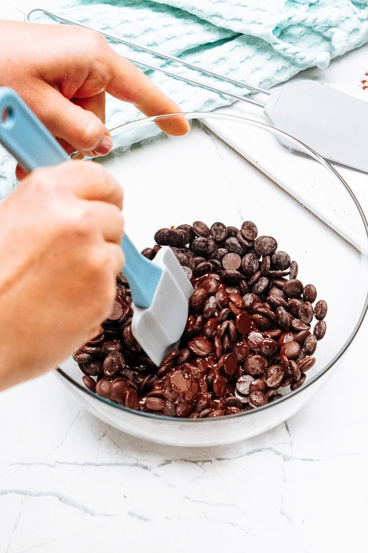 Hands stirring chocolate chips in a glass bowl with a light blue spatula on a pale surface, crafting the perfect chocolate mousse foundation.