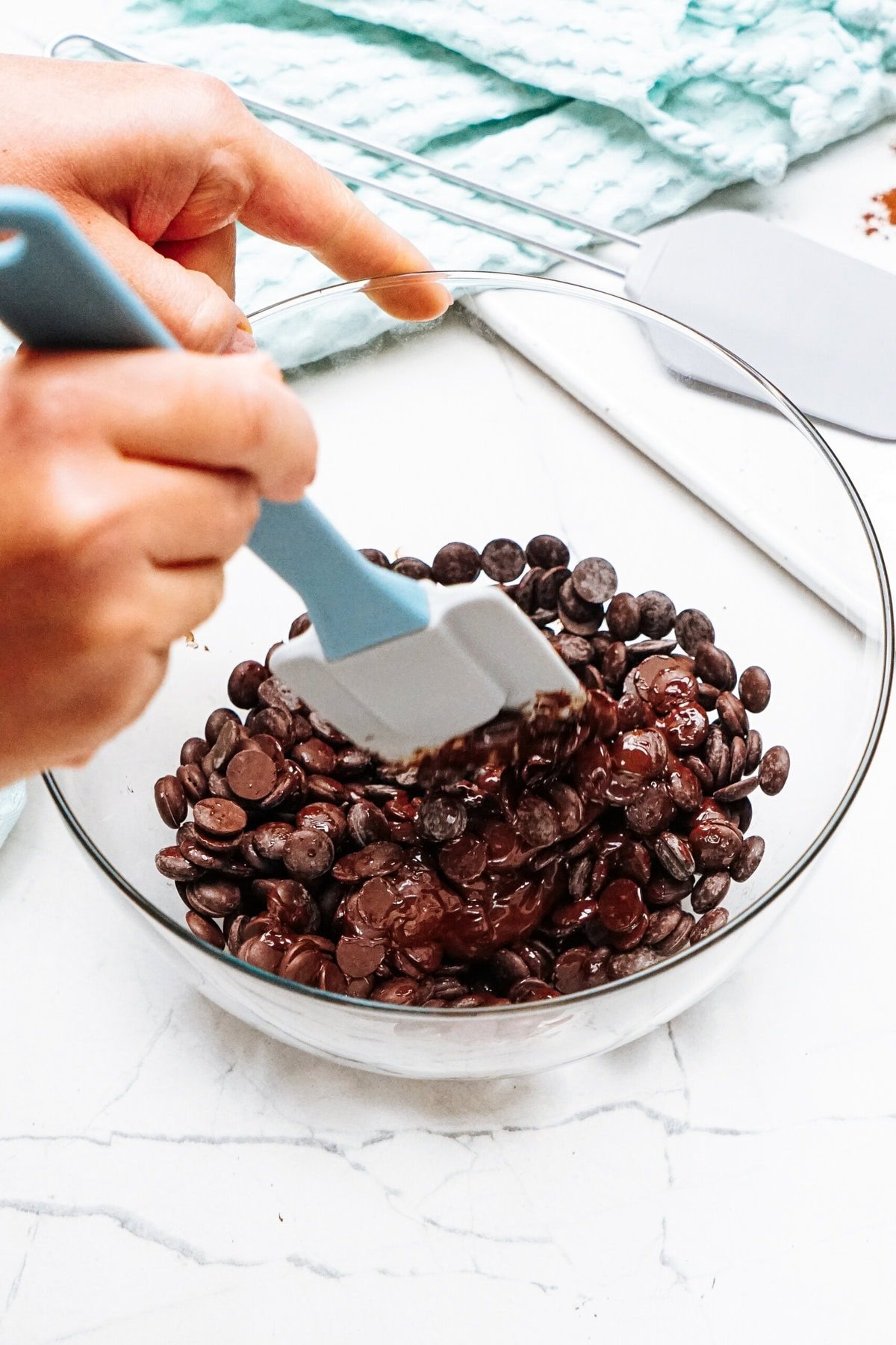 Hands using a blue spatula to stir a bowl of chocolate chips on a marble surface.