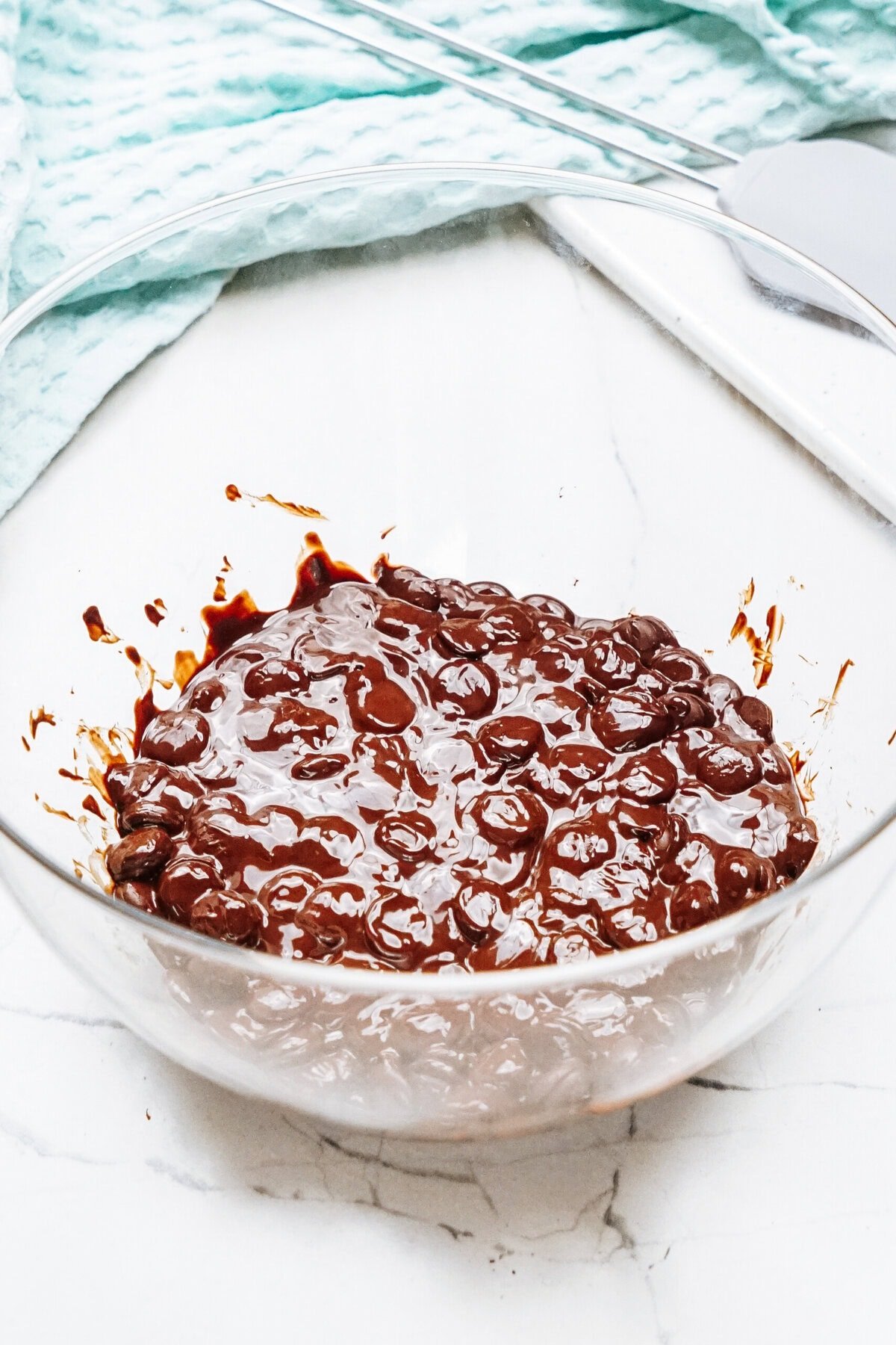 A glass bowl filled with a rich chocolate mousse mixture rests on a marble countertop, accompanied by a towel and metal utensils nearby.