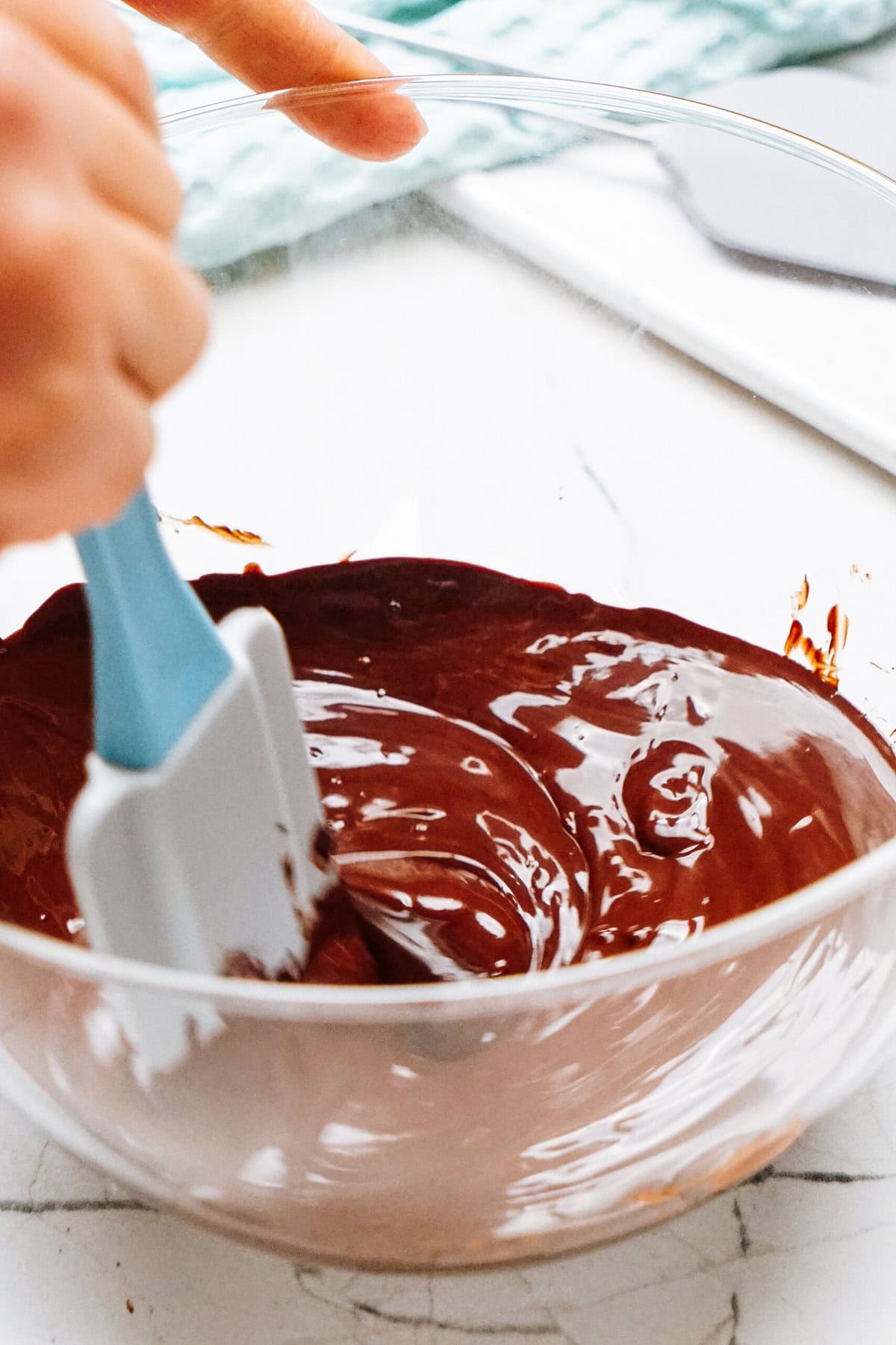 A person stirs melted chocolate destined for a rich chocolate mousse in a clear bowl using a blue spatula.