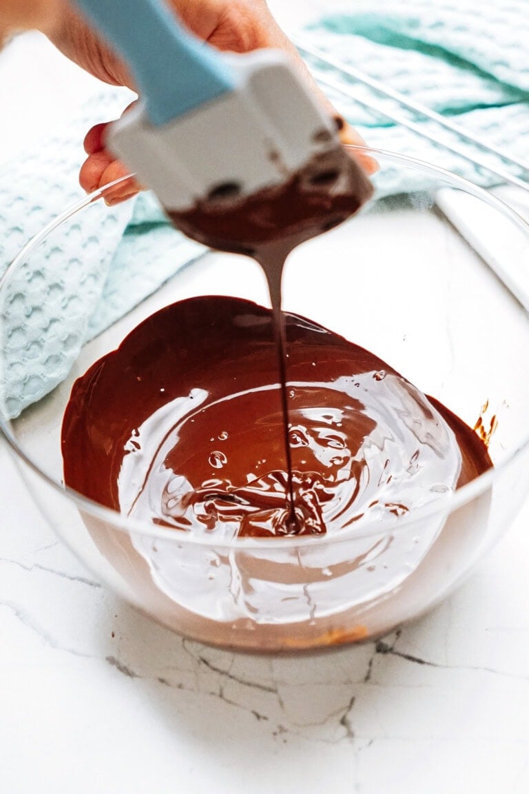 Person stirring melted chocolate in a clear glass bowl with a spatula on a marble surface.