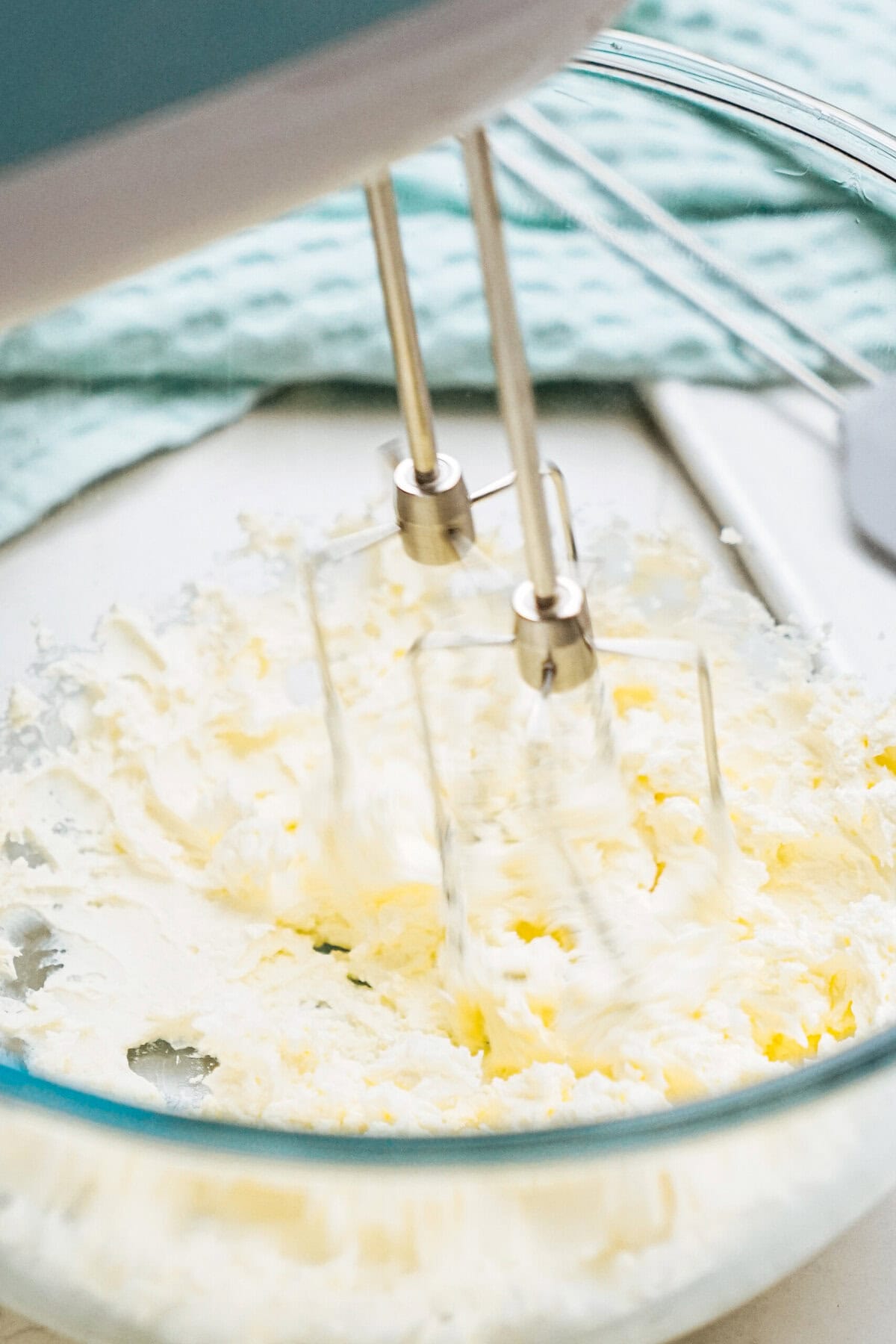 A close-up of a hand mixer with two metal beaters blending cream for chocolate mousse in a glass bowl, set on a light-colored surface.