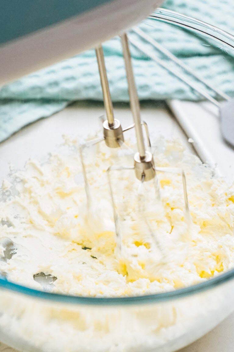 A close-up of a hand mixer whipping butter in a glass bowl.