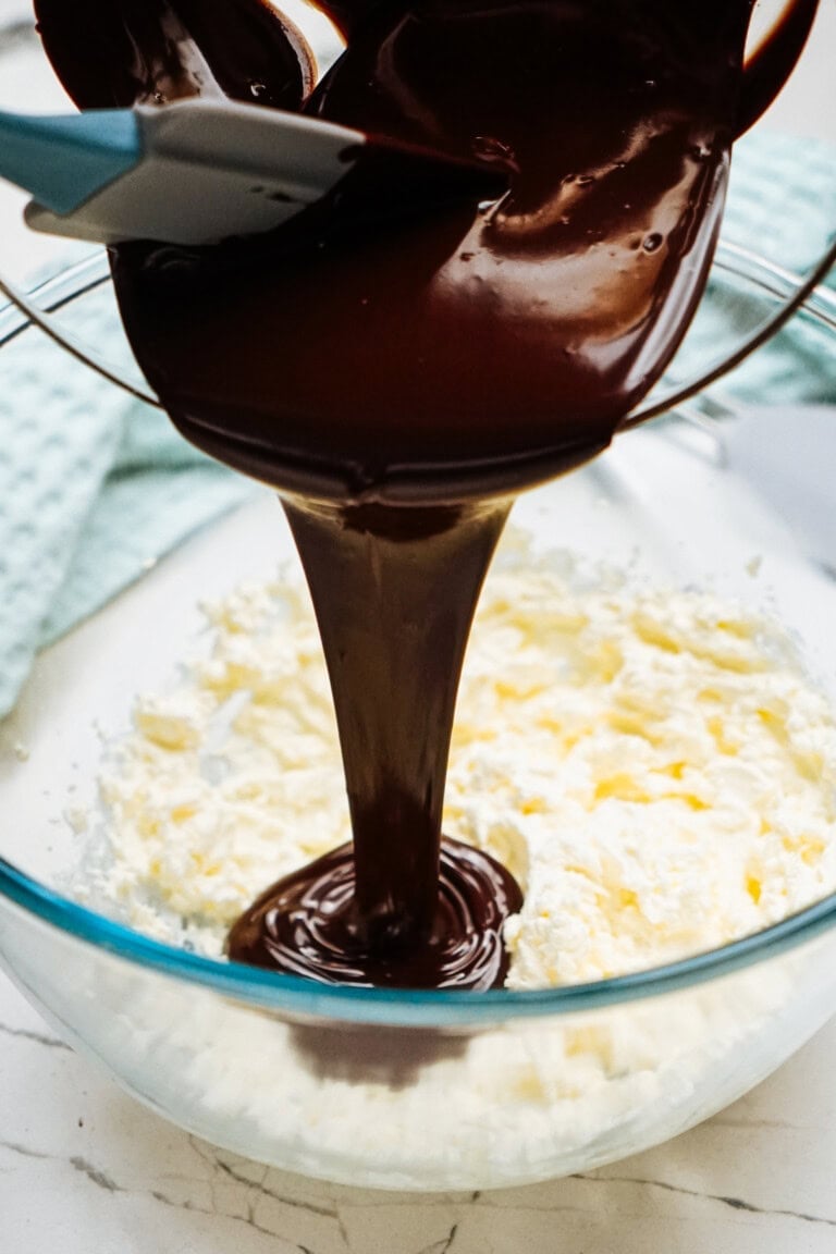 Melted chocolate is being poured into a glass bowl of creamed butter and sugar, beginning the creation of a silky chocolate mousse. A blue spatula is partially visible, ready to fold the mixture into perfection.