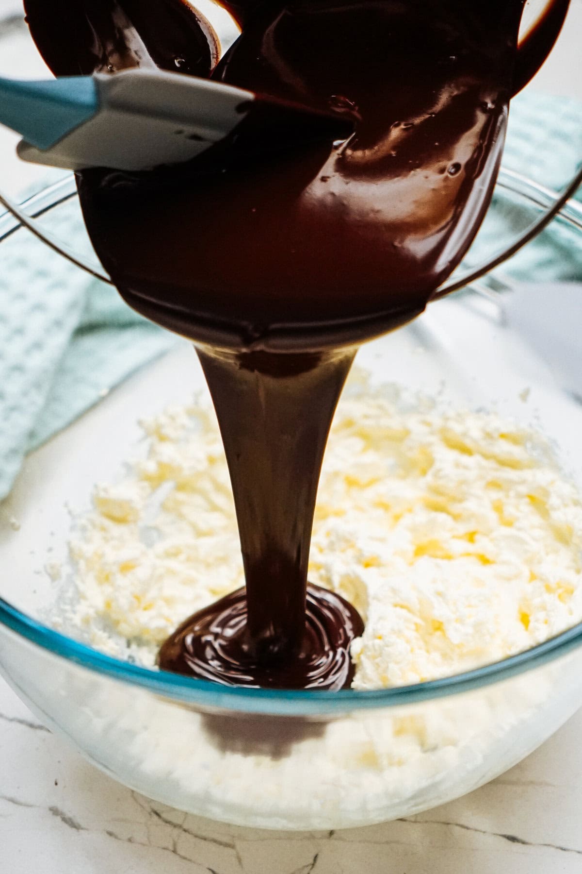 Melted chocolate is being poured from a spatula into a glass bowl of whipped cream.