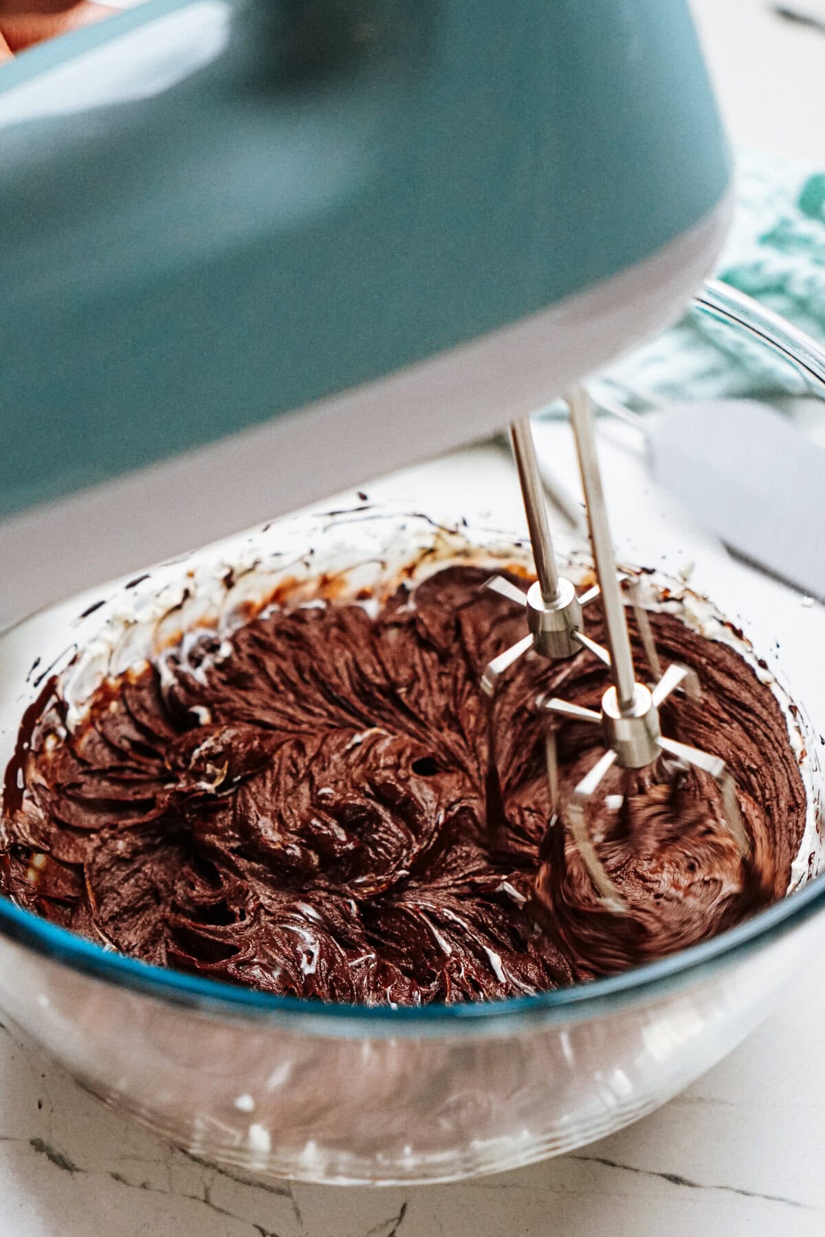 A blue electric mixer blends chocolate batter in a glass bowl on a marble countertop.