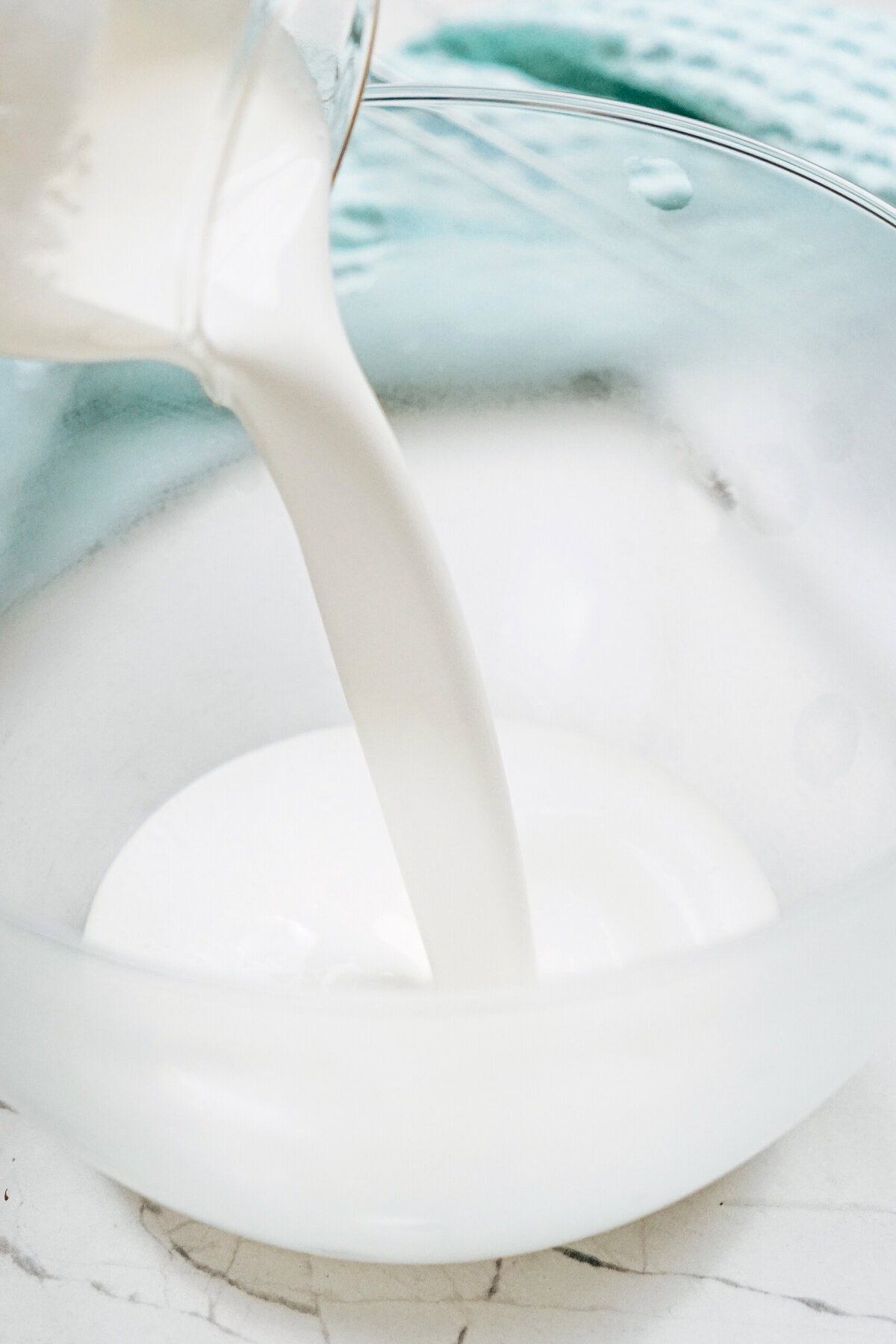 Milk being poured into a clear glass bowl on a white marble surface with a blue cloth in the background.