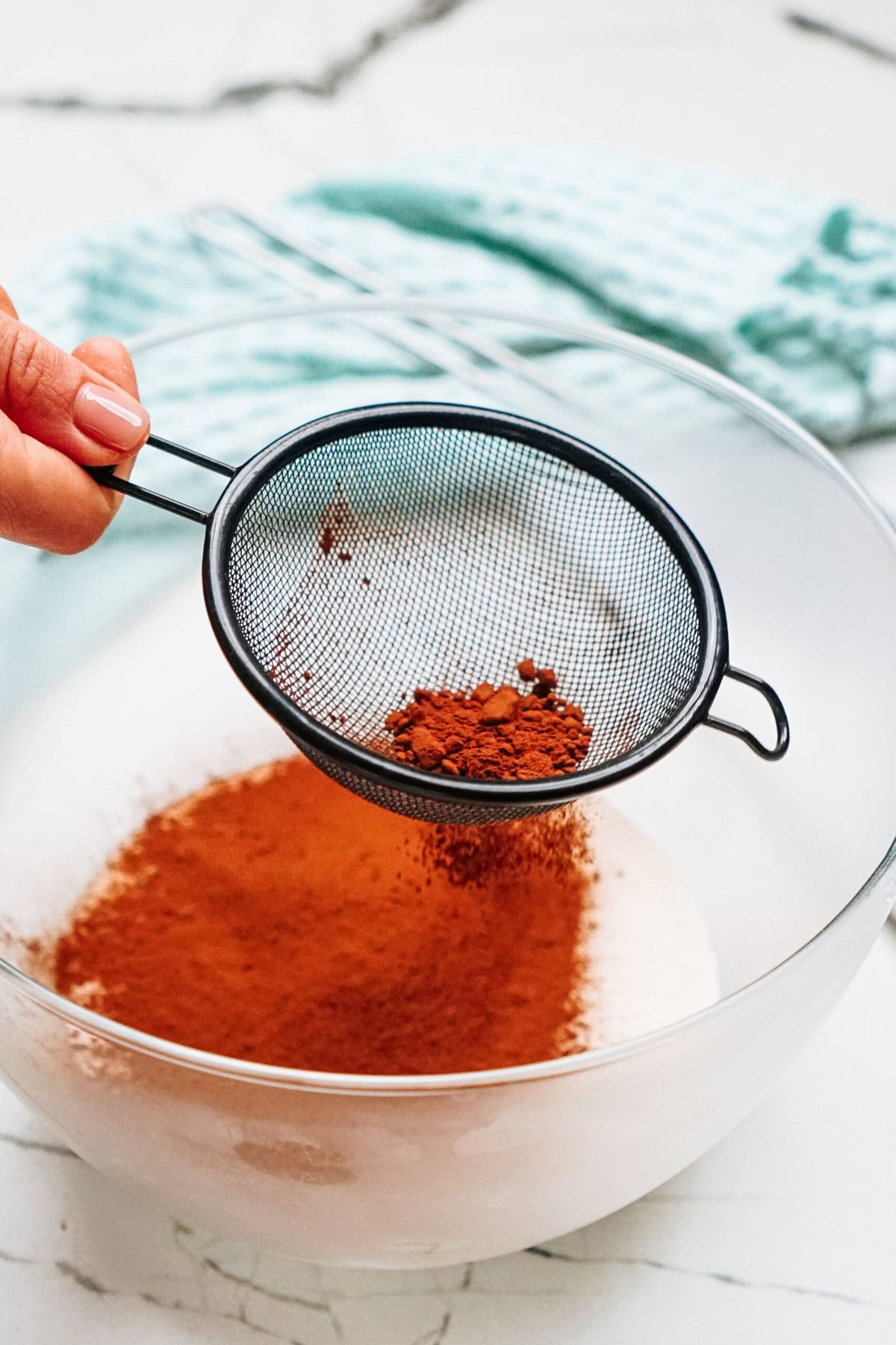 A hand sifts cocoa powder into a glass bowl on a marble countertop. A light blue cloth is in the background.