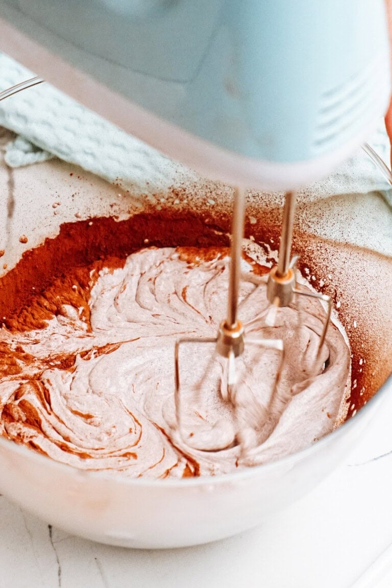 An electric mixer whirls the cocoa powder into a creamy chocolate mousse in a glass bowl on the white countertop.