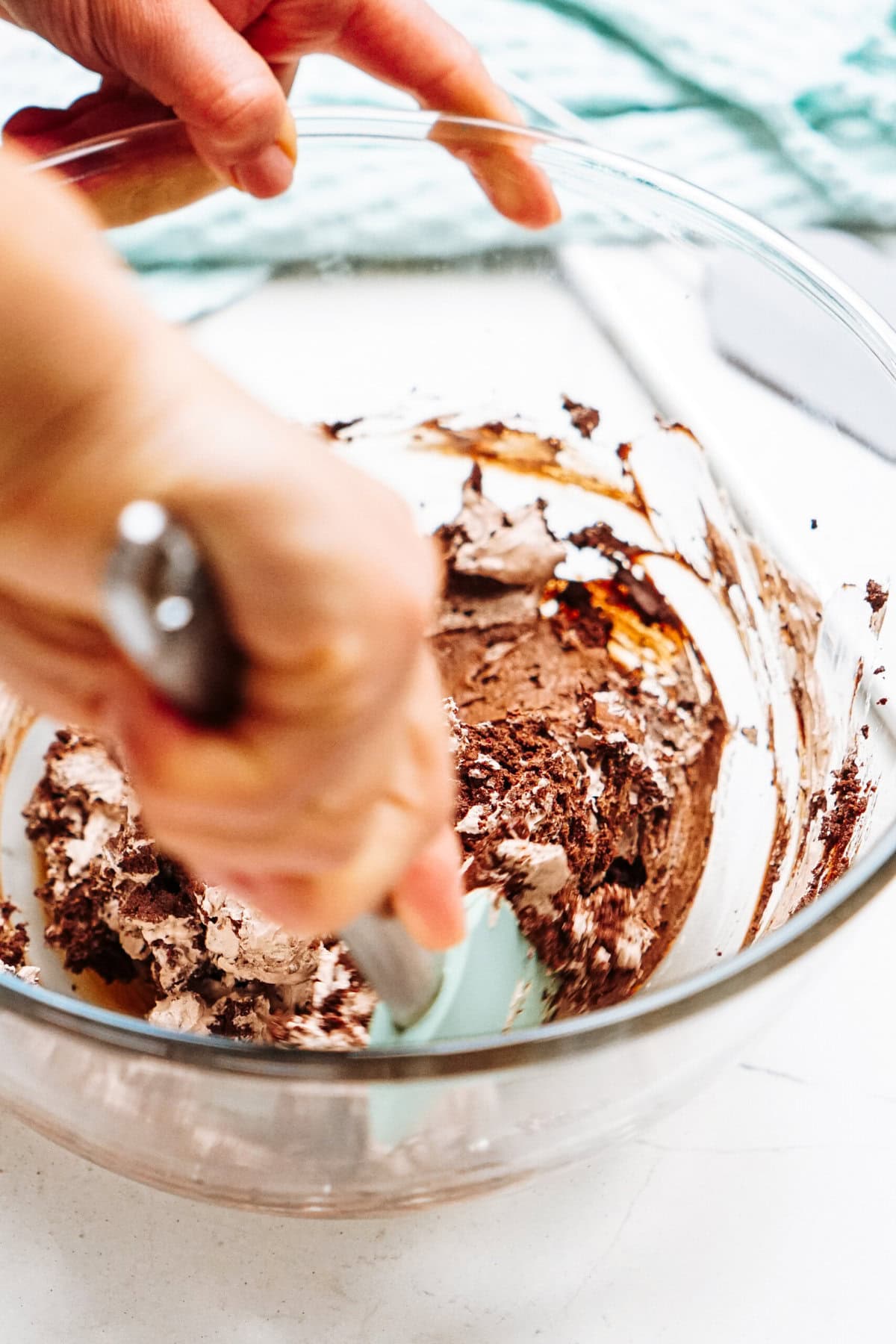 A person mixes chocolate mousse batter in a clear glass bowl with a spatula.