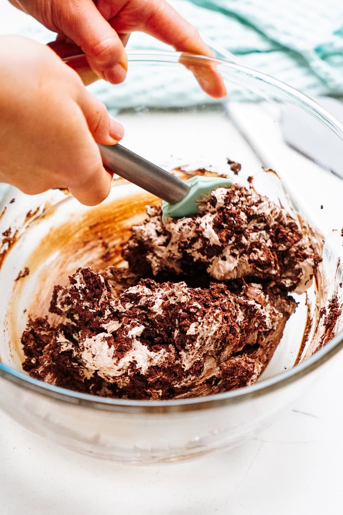 Hands mixing chocolate batter in a glass bowl with a spatula.