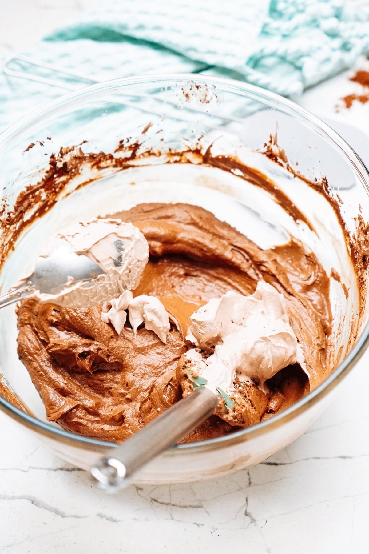 A glass bowl with chocolate batter being mixed by a metal spoon on a marble surface, next to a blue cloth.