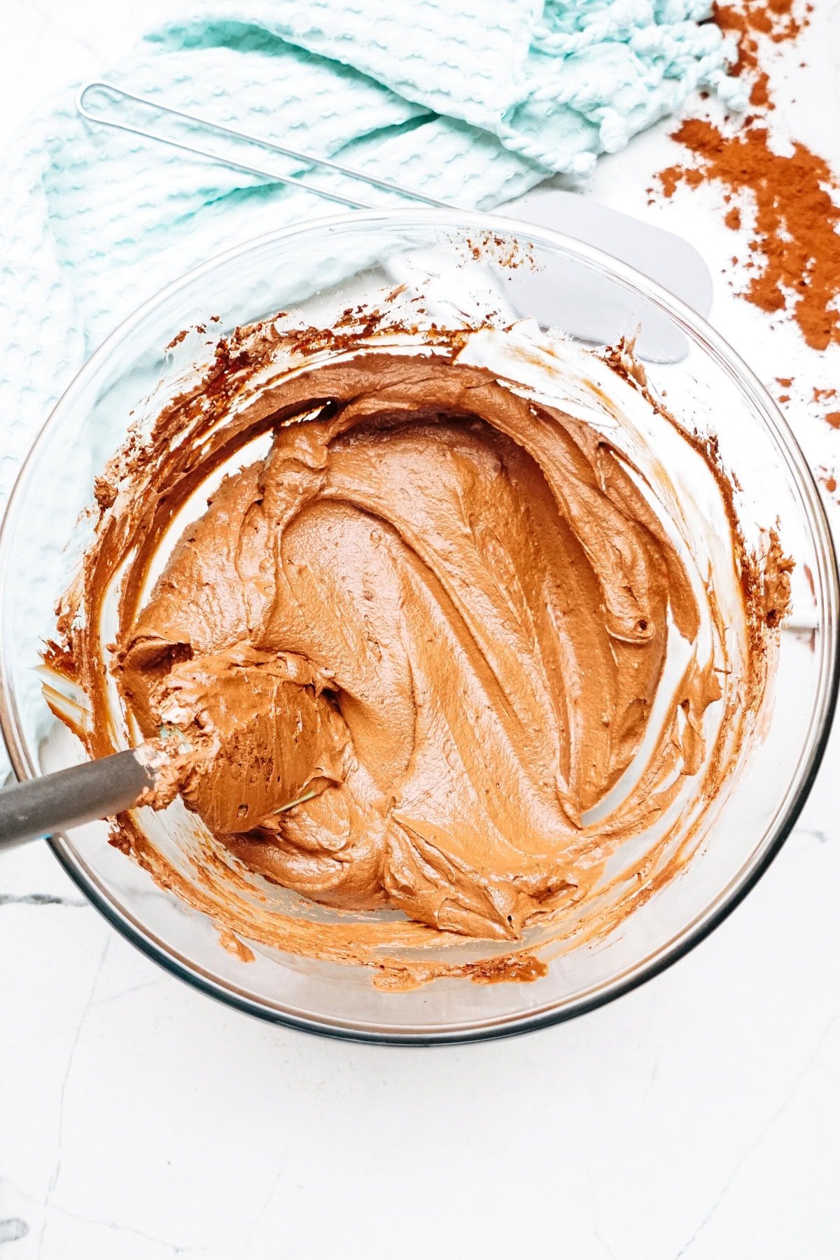 A glass bowl filled with creamy chocolate mousse frosting, with a spatula resting inside. A light blue towel and cocoa powder are nearby on a white surface.