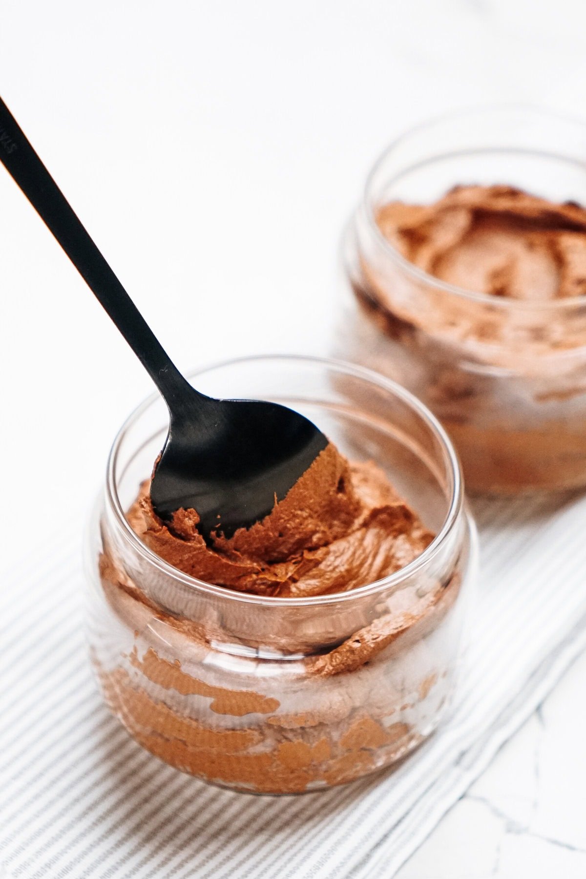 Two glass jars brimming with rich, creamy chocolate mousse sit invitingly on a striped cloth. A black spoon gracefully scoops the decadent mousse from one jar, promising a moment of pure indulgence.