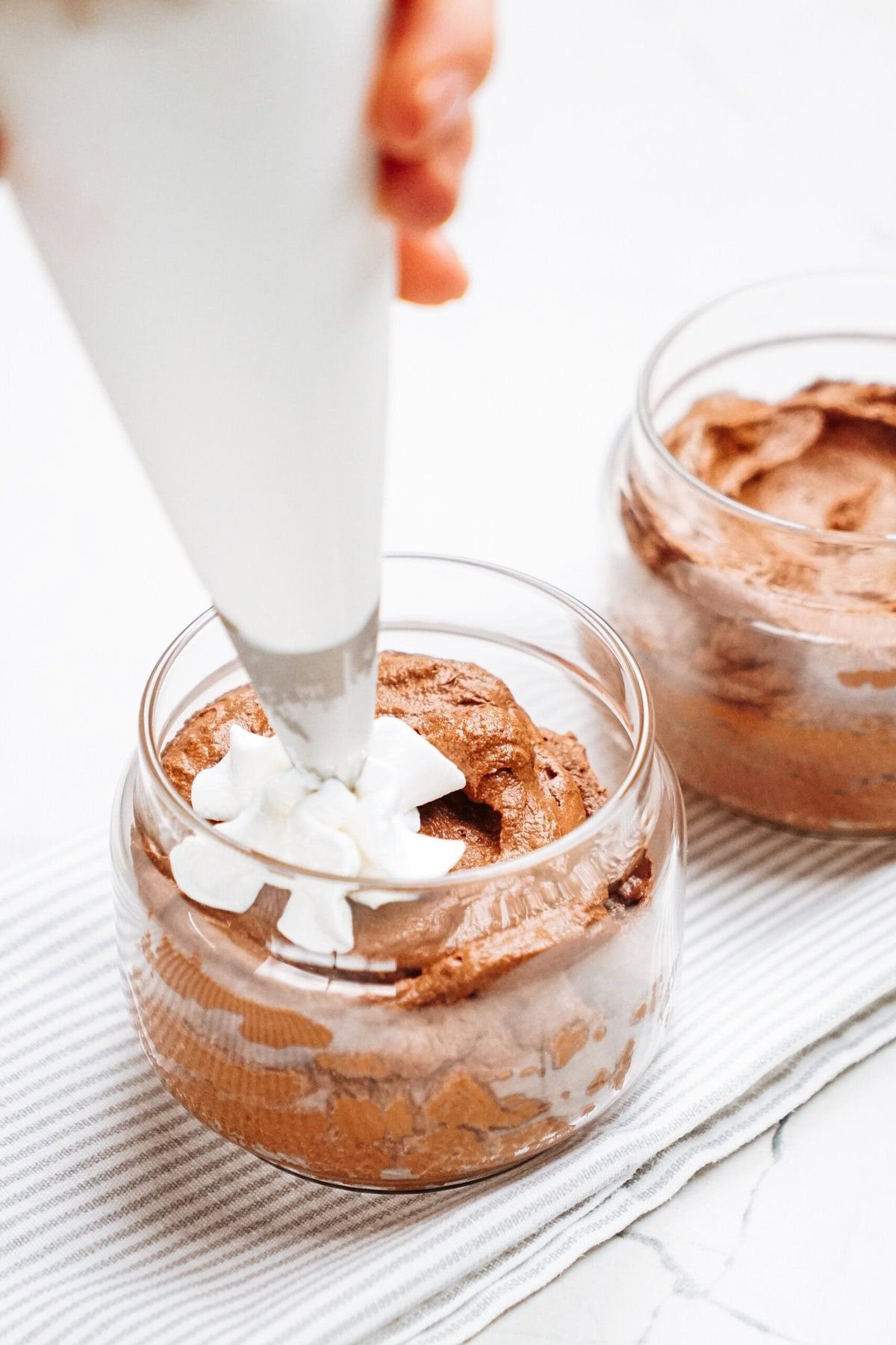 Delicately piping whipped cream onto the luscious chocolate mousse nestled in a glass container, all set on a charming striped cloth.