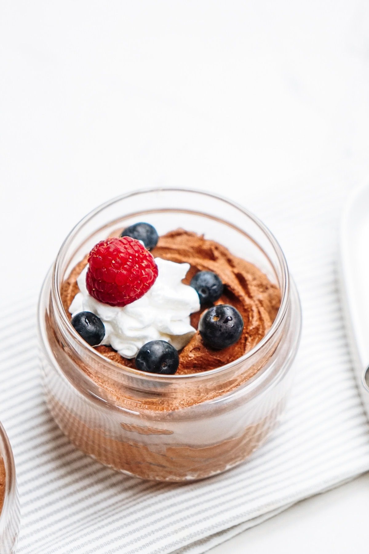 A jar of decadent chocolate mousse is elegantly topped with a swirl of whipped cream, a fresh raspberry, and vibrant blueberries, all set on a striped cloth.