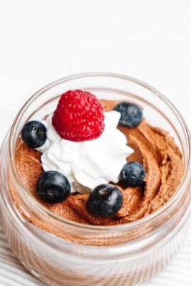 Indulge in a delightful dessert in a jar, featuring rich chocolate mousse topped with a swirl of whipped cream, crowned with a juicy raspberry and fresh blueberries.