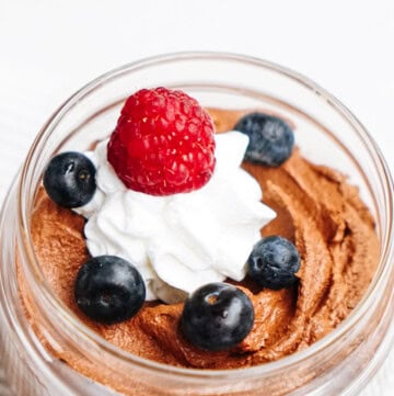 Indulge in a delightful dessert in a jar, featuring rich chocolate mousse topped with a swirl of whipped cream, crowned with a juicy raspberry and fresh blueberries.