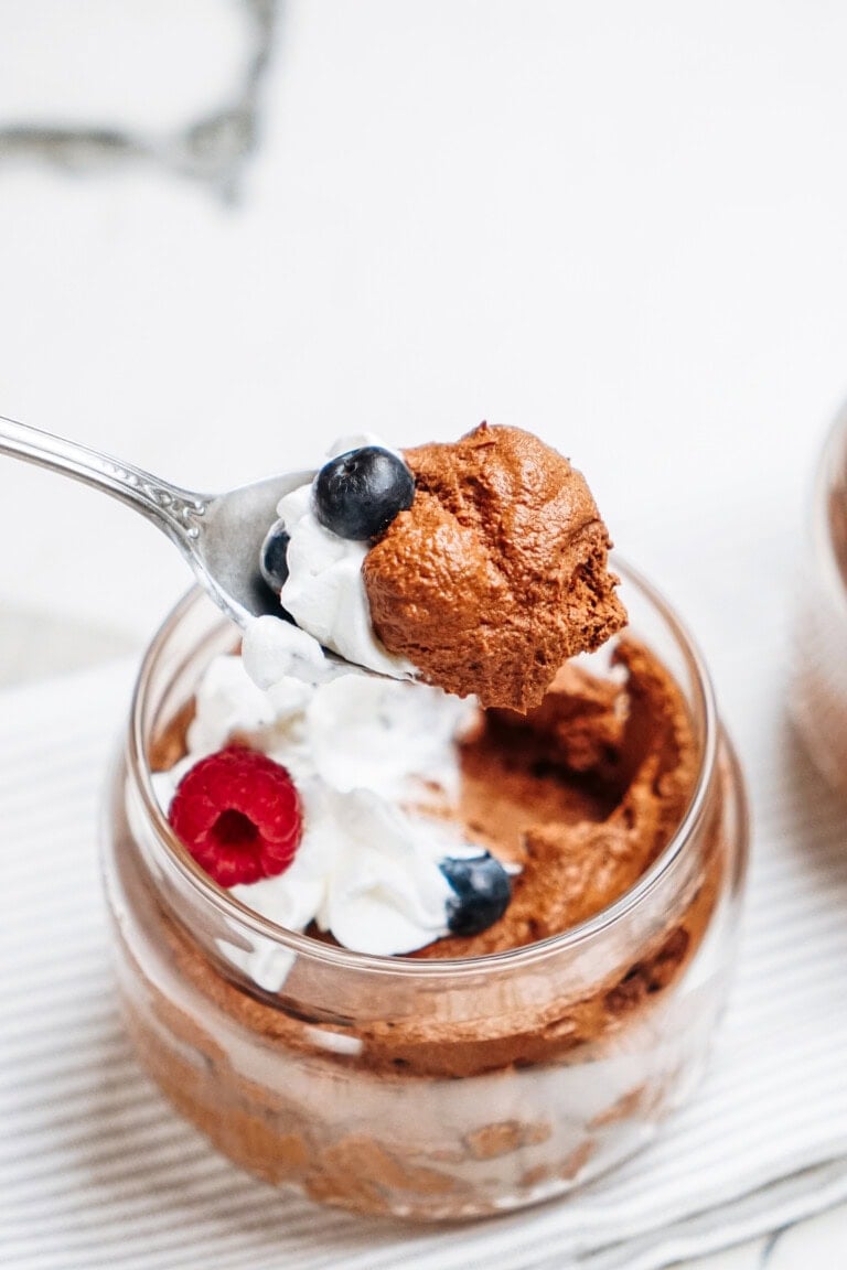 A spoonful of rich chocolate mousse with whipped cream, topped with a blueberry and a raspberry, sits temptingly. In the background, a glass jar brims with more decadent mousse and fresh fruit.